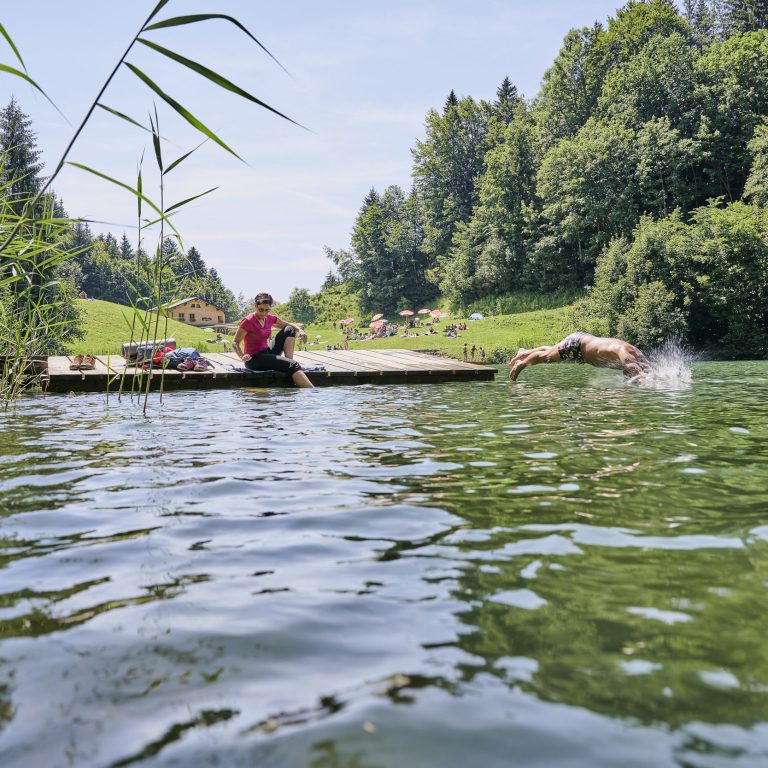 Abkühlung im Seewaldsee im Biosphärenpark Großes Walsertal, Eine Person sitzt am STeg und hält die Füße ins Wasser, Eine Person springt mit einem Kopfsprung ins Wasser.