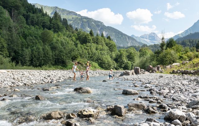 Eine Familie mit Kindern spielt an der Bregenzer Ach, im Hintergrund die grüne Natur und Berglandschaft des Bregenzerwaldes