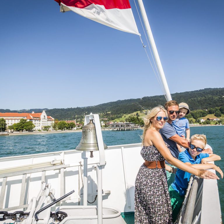 Eine Familie steht sommerlich gekleidet am Bug eines Schiffes der Weißen Flotte der Vorarlberg Lines - Bodenseeschifffahrt. Im Hintergrund ist das Seehotel Am Kaiserstrand in Lochau zu sehen. Die Fahne weht im Fahrtwind.