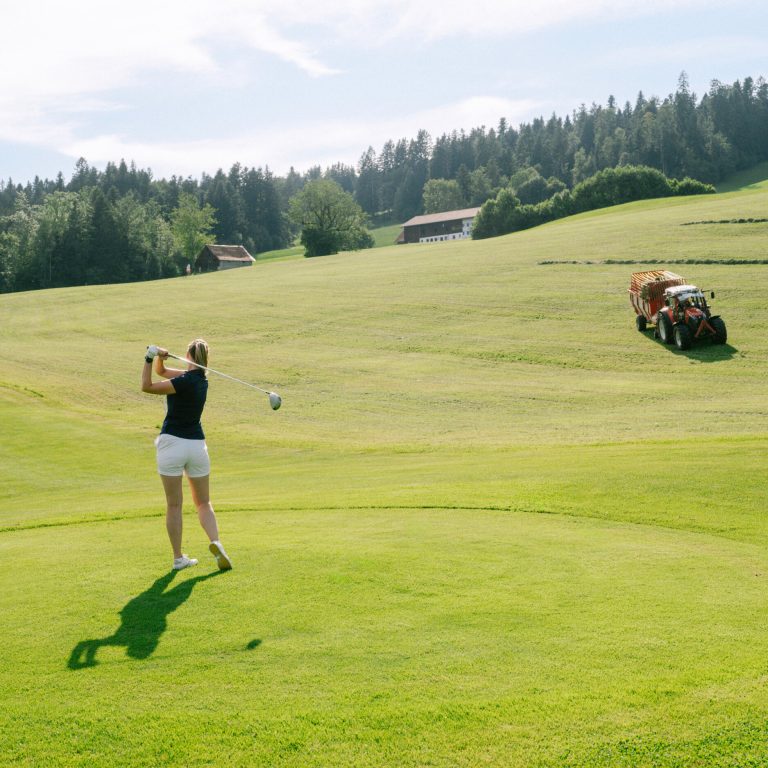 Eine Frau schwingt ihren Golfschläger am Golfplatz im Bregenzerwald, im Hintergrund fährt ein Traktor. 