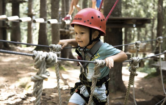 Ein Kind mit Kletterhelm und Klettergurt klettert gesichert am Seil über eine Seilbrücke im Waldseilgarten Damüls.