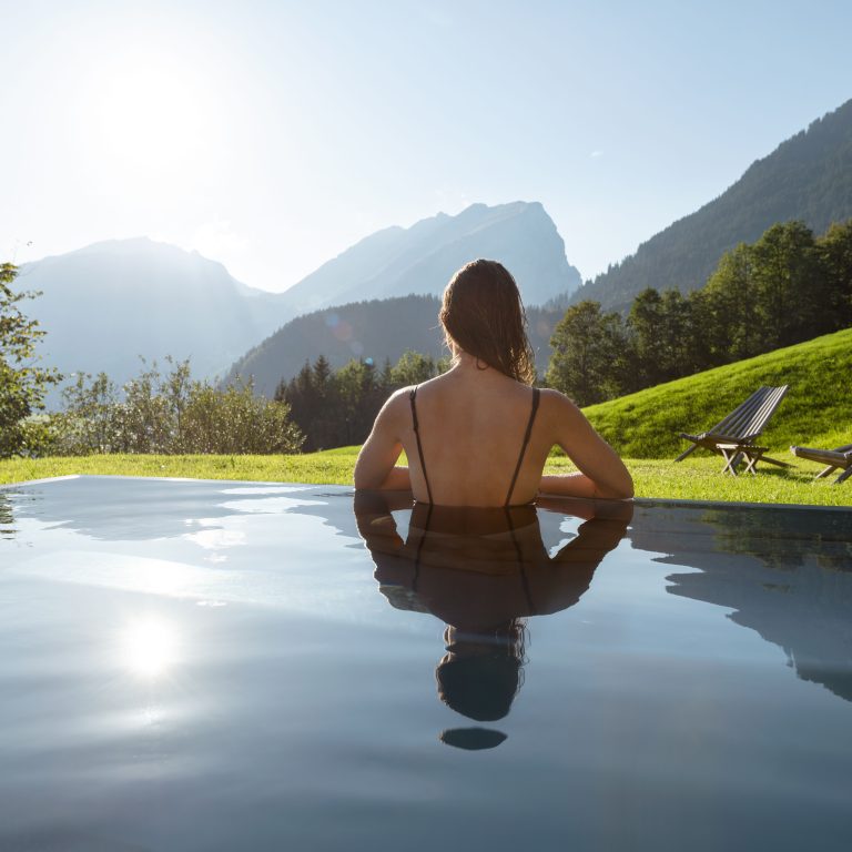 Frau in Rückenansicht, steht im Wasser am Rand eines Aussenpools und blickt auf die grüne Landschaft im Bregenzerwald hinaus