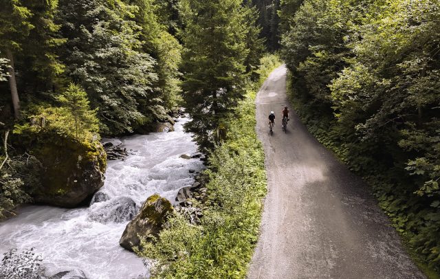 Zwei Radfahrende auf einer schattigen Straße entlang eines Gebirgsbachs in Vorarlberg