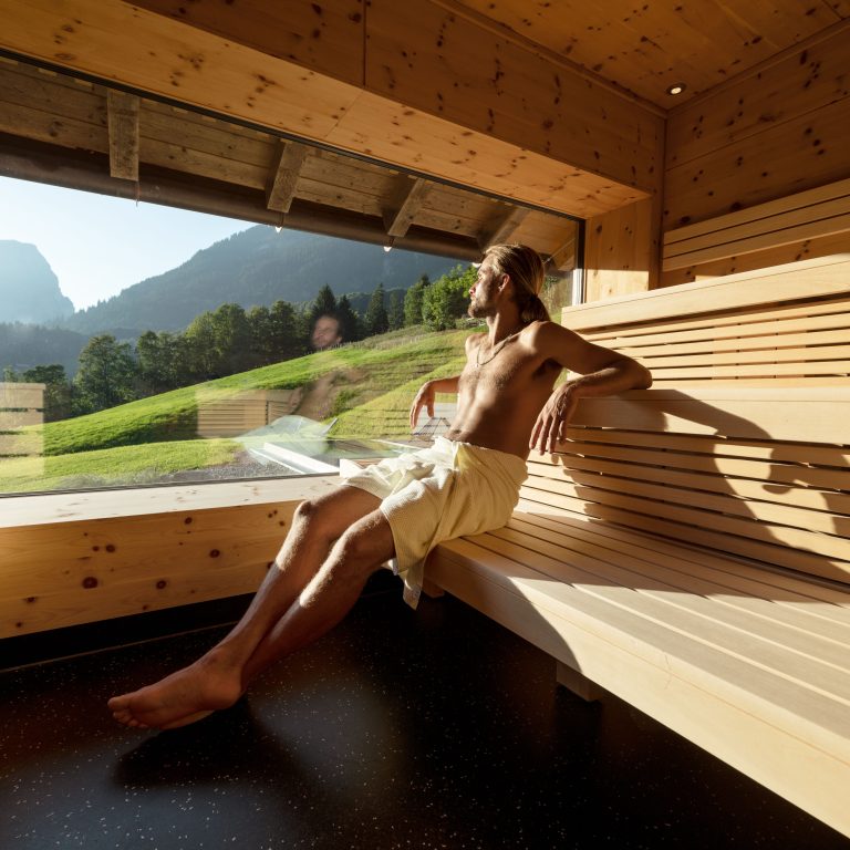 Junger Mann sitzt in der Sauna und blickt aus dem Panoramafenster in die grüne Landschaft im Bregenzerwald